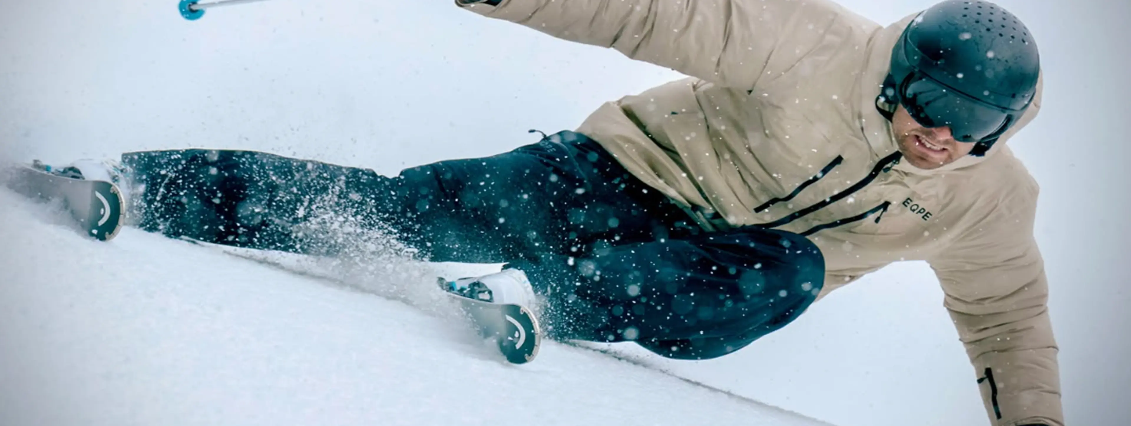 A skier carves through the snow, wearing a beige jacket and black helmet, creating a spray of snow and showcasing dynamic motion.
