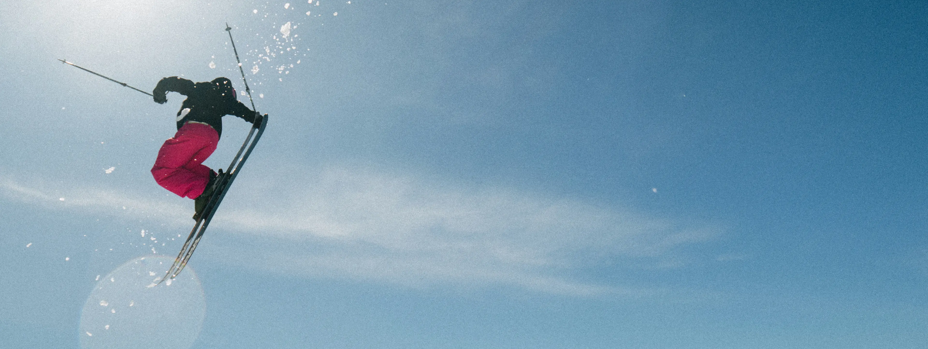 A skier in mid-air performing a jump against a clear blue sky, snow particles scattering around.
