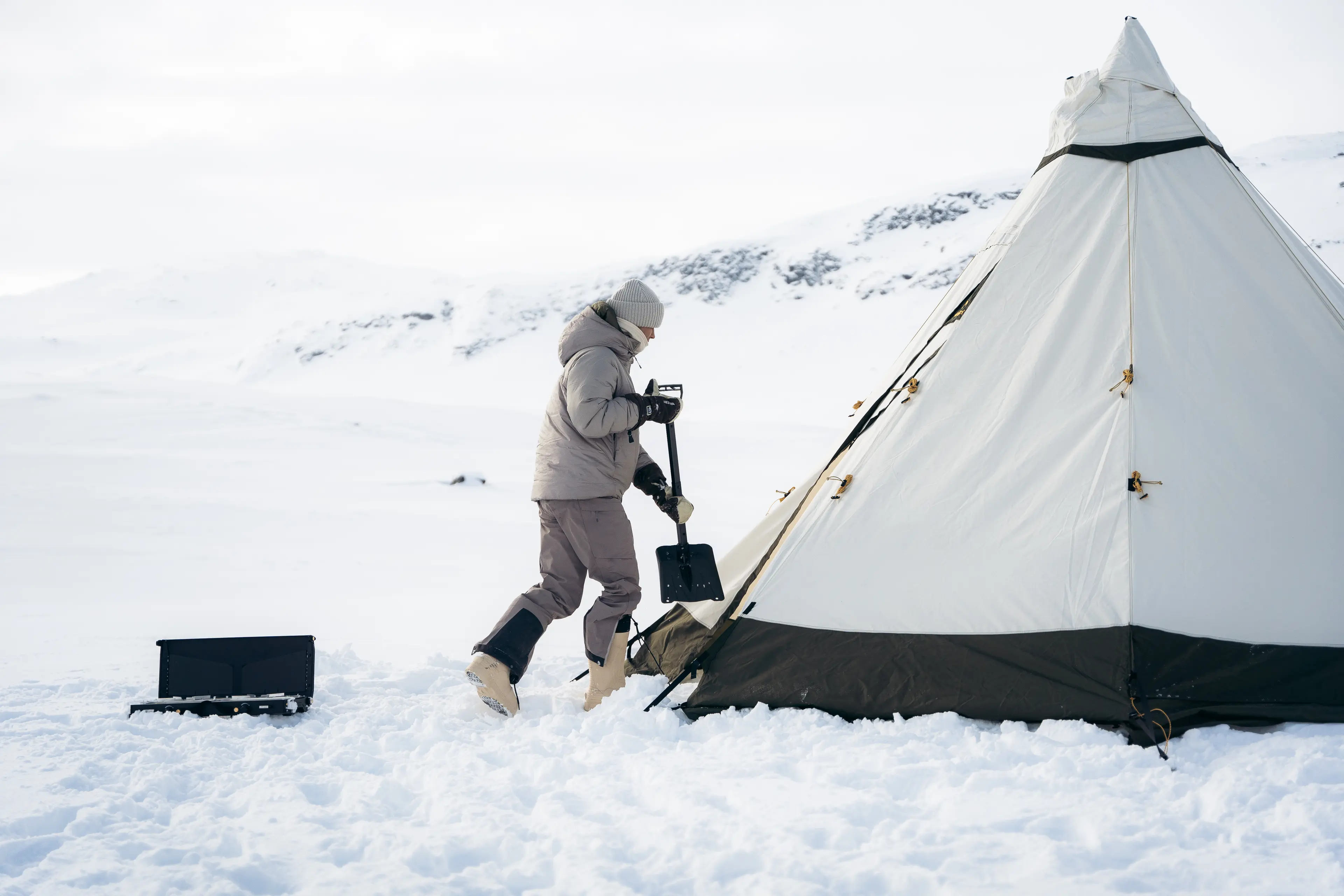 Person in winter gear shovels snow near a large, beige tent in a snowy landscape with mountains in the background.