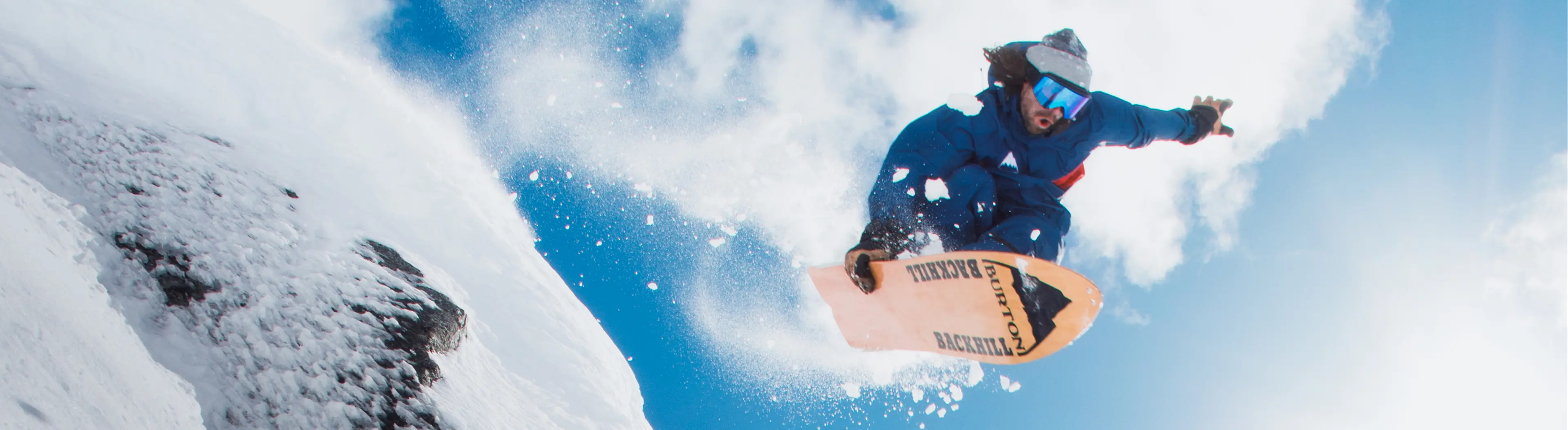 A snowboarder in blue gear is captured mid-air against a bright sky, kicking up snow as they perform a trick on a snowy slope.