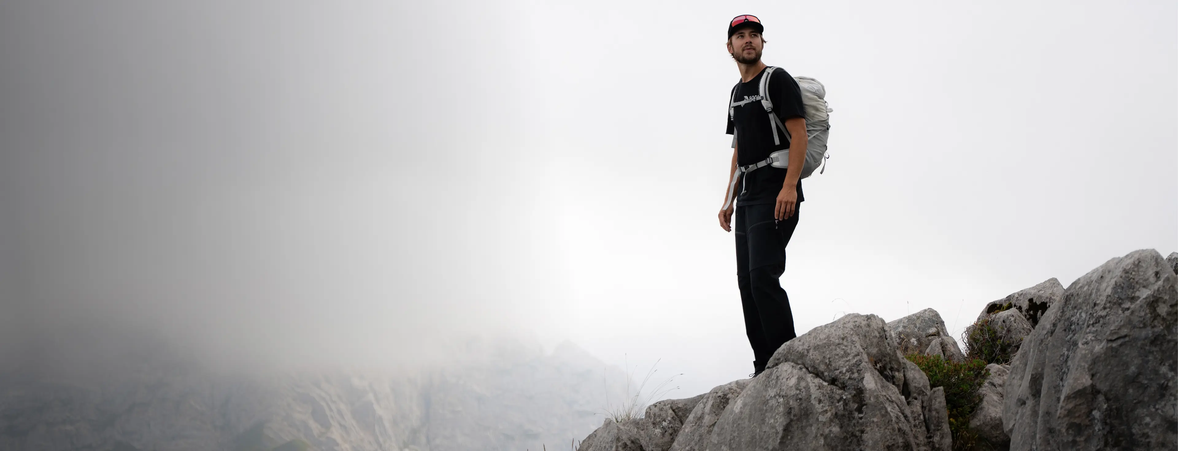 A person stands on a rocky mountain peak with a backpack, looking into the distance. The background is misty and overcast.