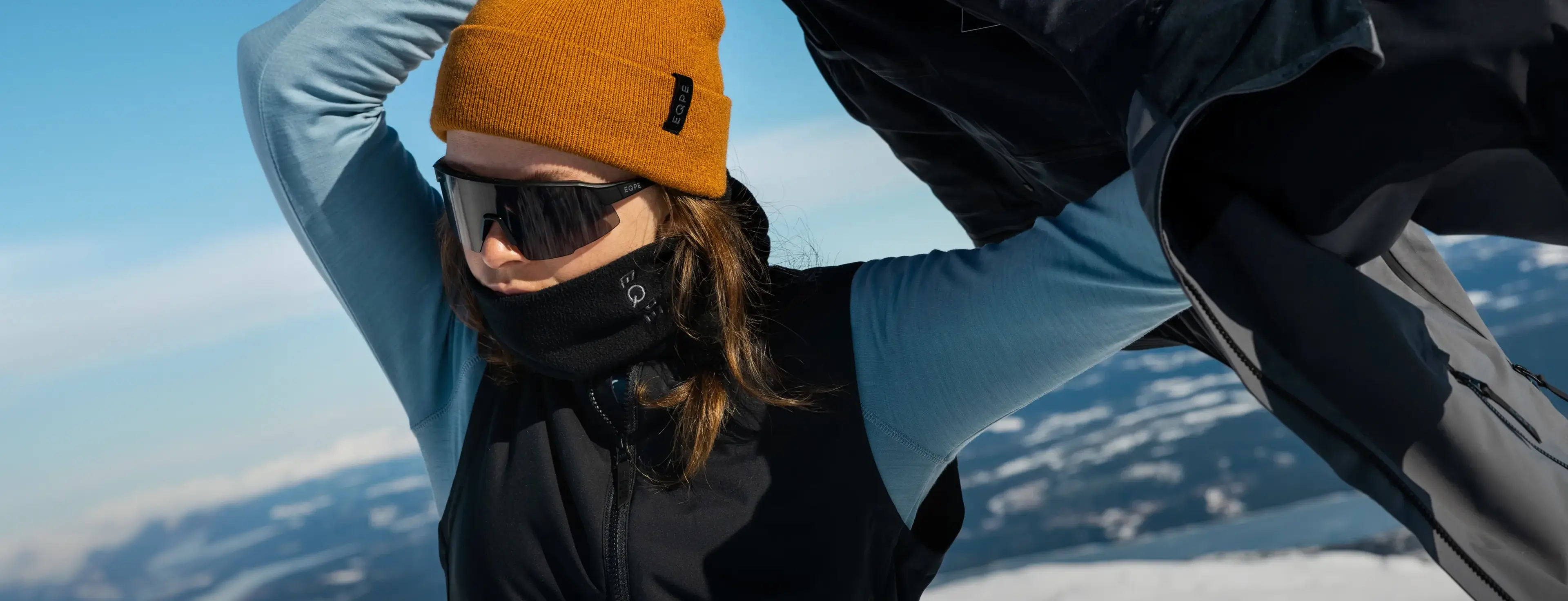 A person in a mustard beanie and sunglasses adjusts their jacket on a snowy mountain, with a clear blue sky in the background.