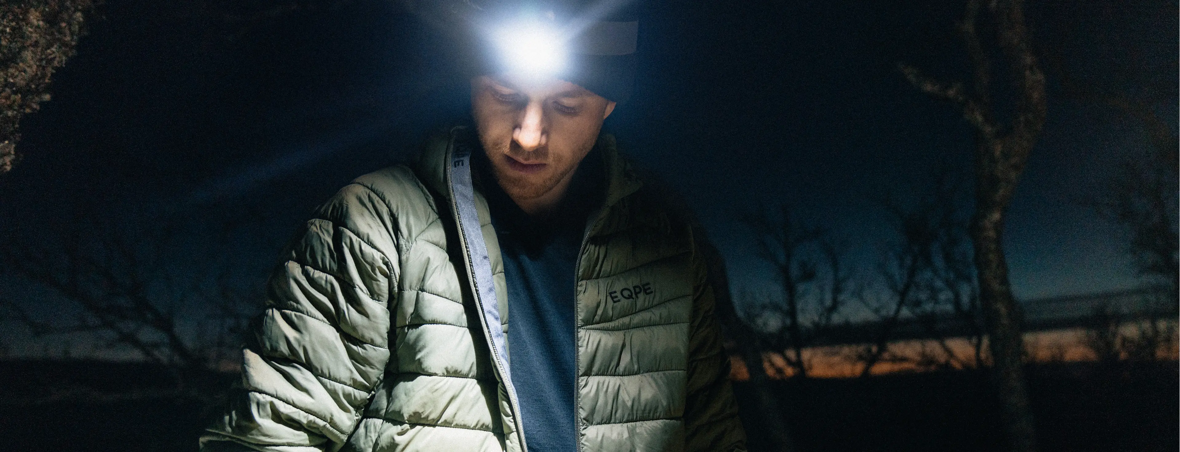A person wearing a headlamp and a padded jacket stands outdoors in the dark, with a dim twilight sky in the background.