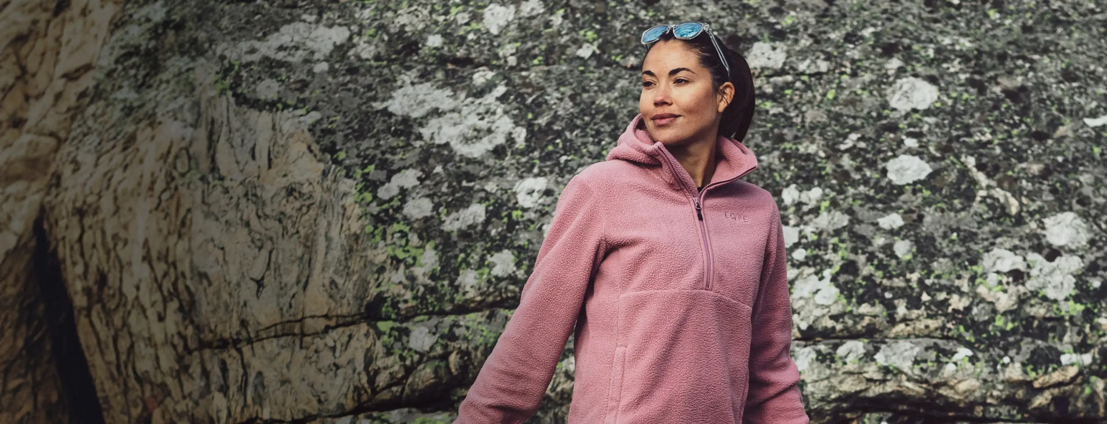 A person in a pink fleece jacket stands confidently in front of a textured rock formation, with sunglasses resting on their head.