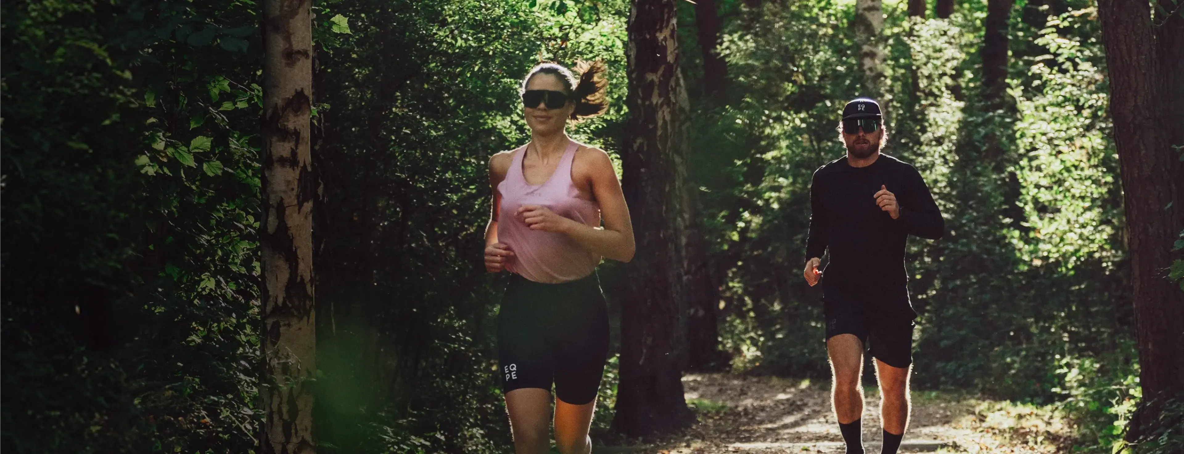 Two people running on a forest trail, one wearing a pink top and the other in black, surrounded by lush greenery.