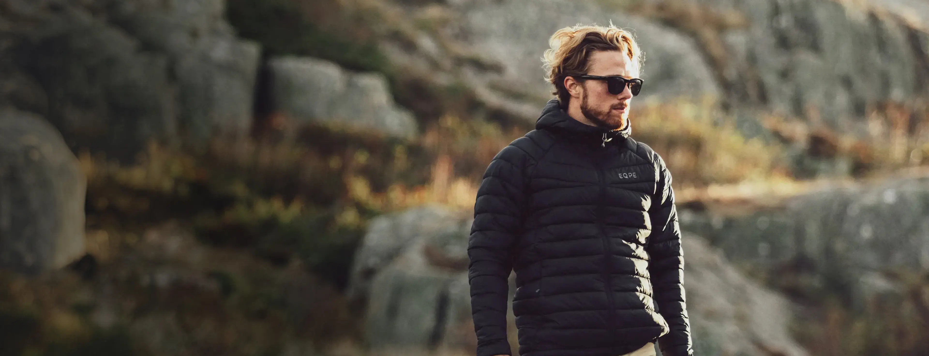 A man with sunglasses and a black puffer jacket walks outdoors, with a rocky landscape in the background.