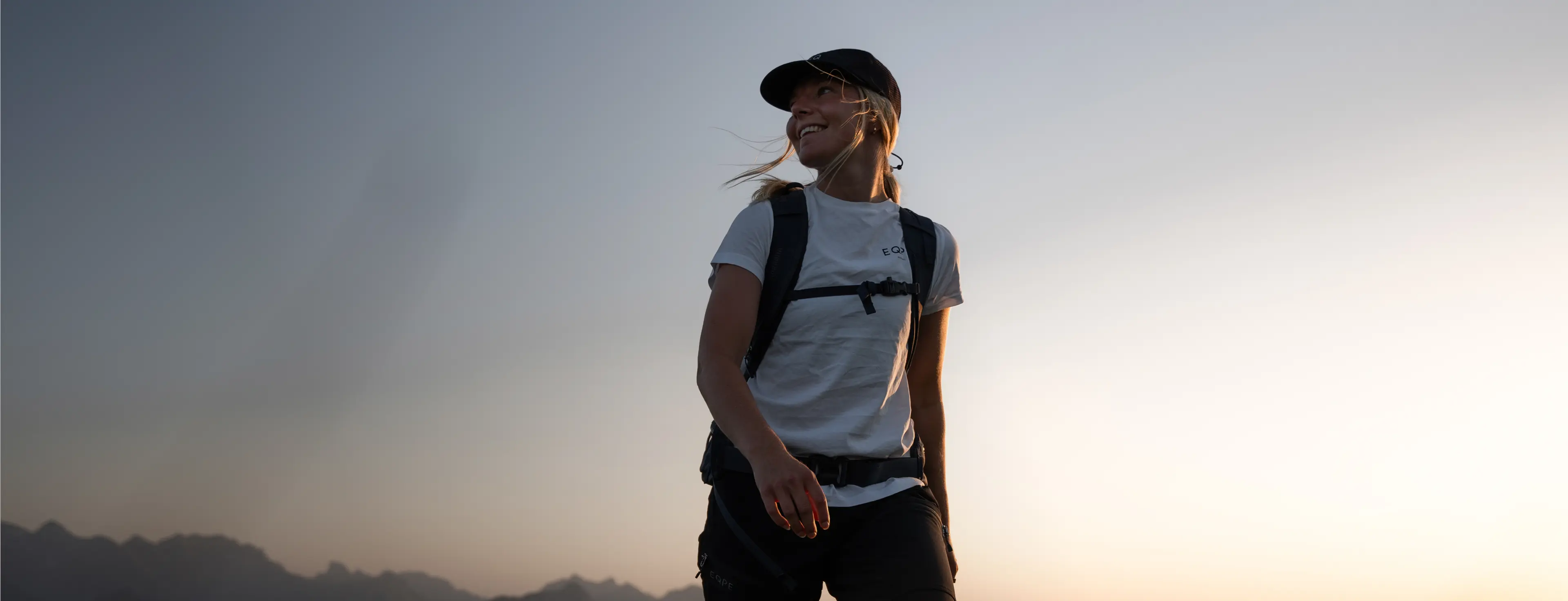A person wearing a cap and backpack stands in front of a dusky sky, smiling and looking to the side, with mountain silhouettes in the background.