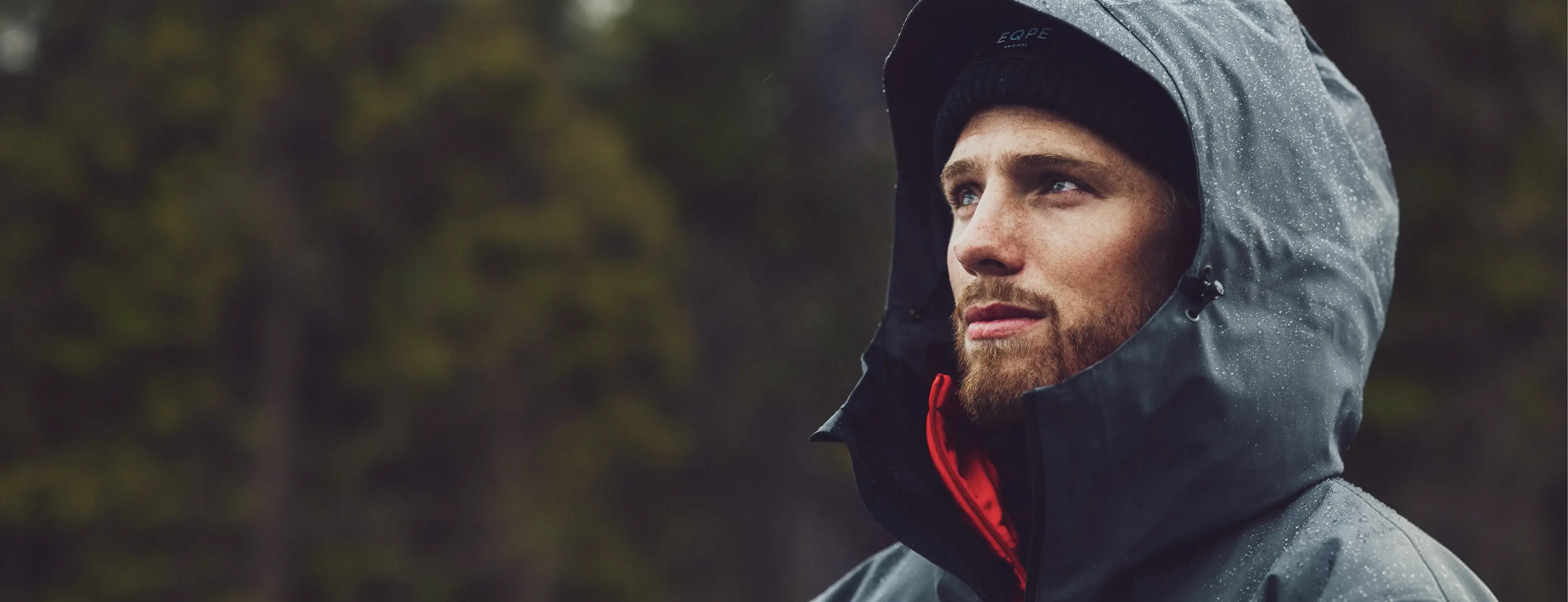 Man wearing a hooded jacket with raindrops looks thoughtfully into the distance.