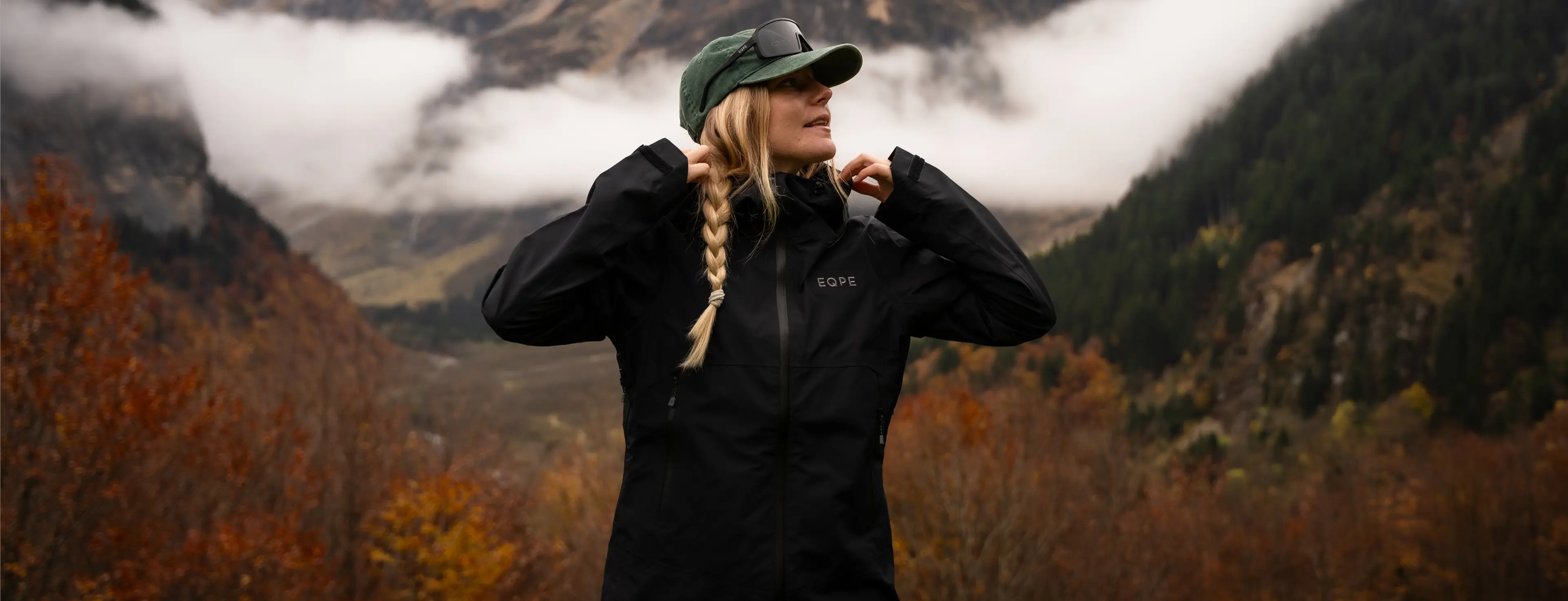 A person with a braid and cap adjusts their jacket, standing in a foggy mountainous landscape with autumn foliage.
