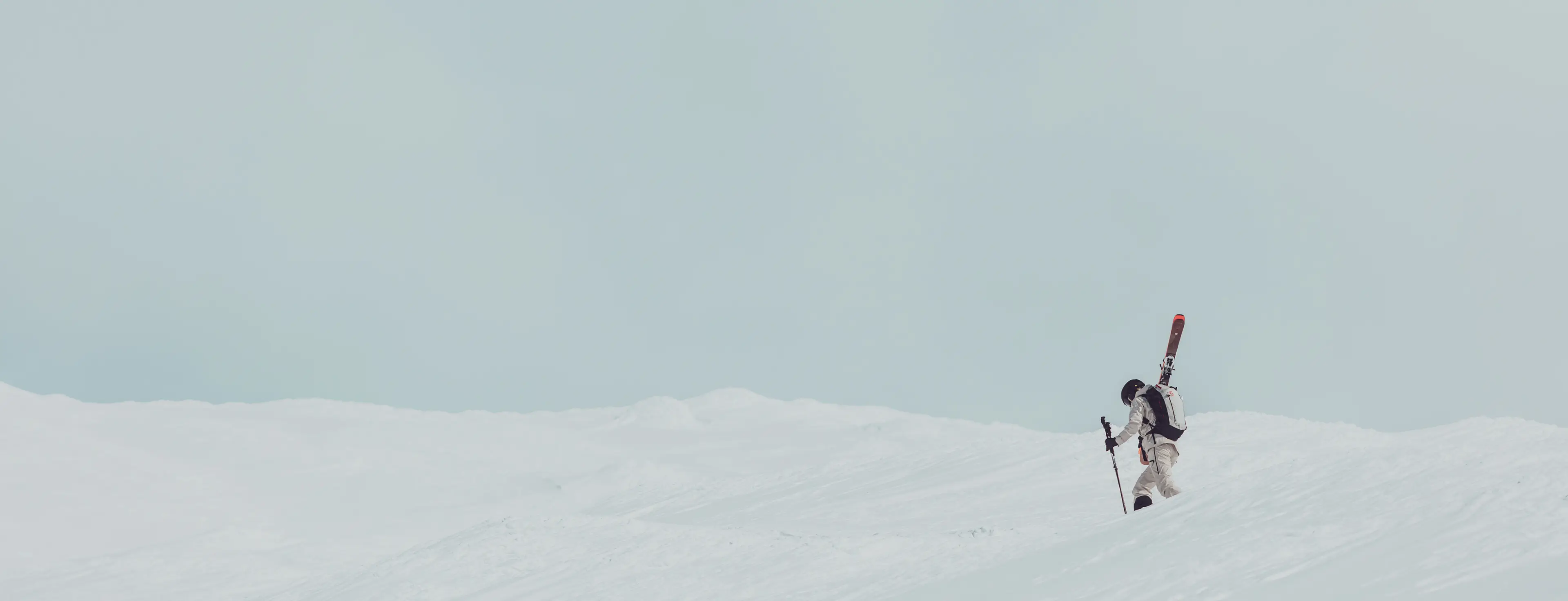 A skier in ski gear climbs a snowy slope under a pale blue sky.