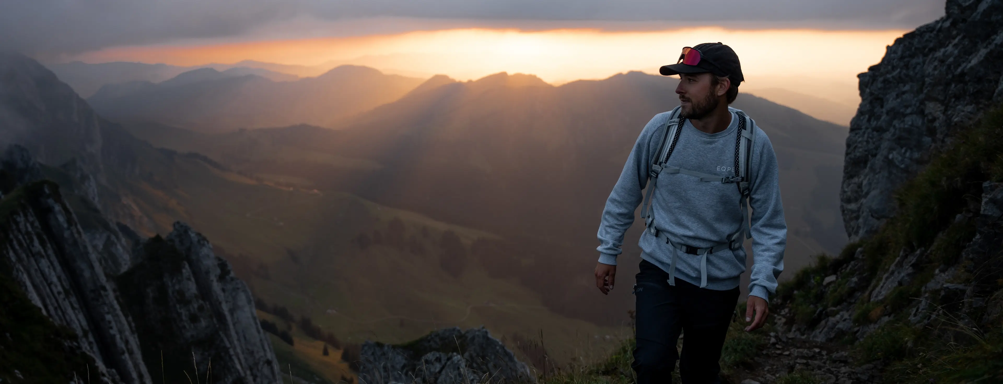 A man hiking on a rocky mountain path at sunset, with sweeping views of the valley and distant peaks illuminated by warm sunlight.