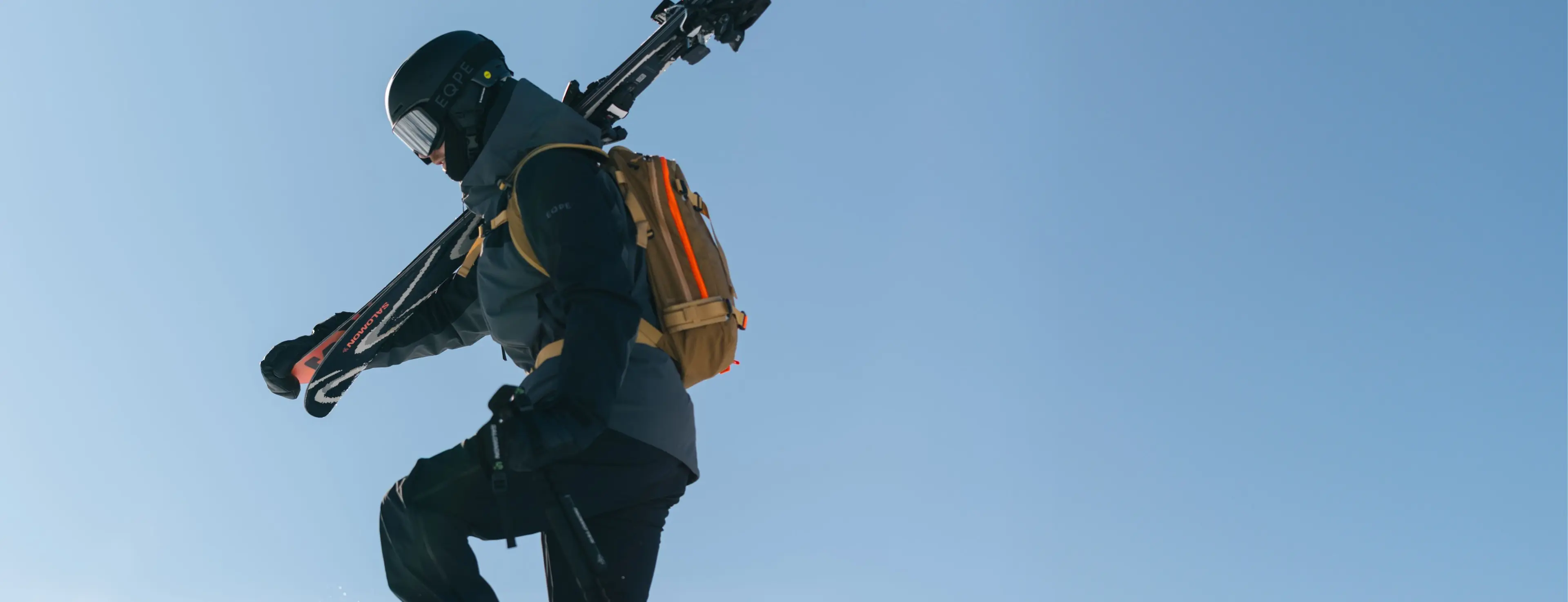 A skier in a helmet carries skis over the shoulder while walking on snow, with a bright orange backpack against a clear blue sky.