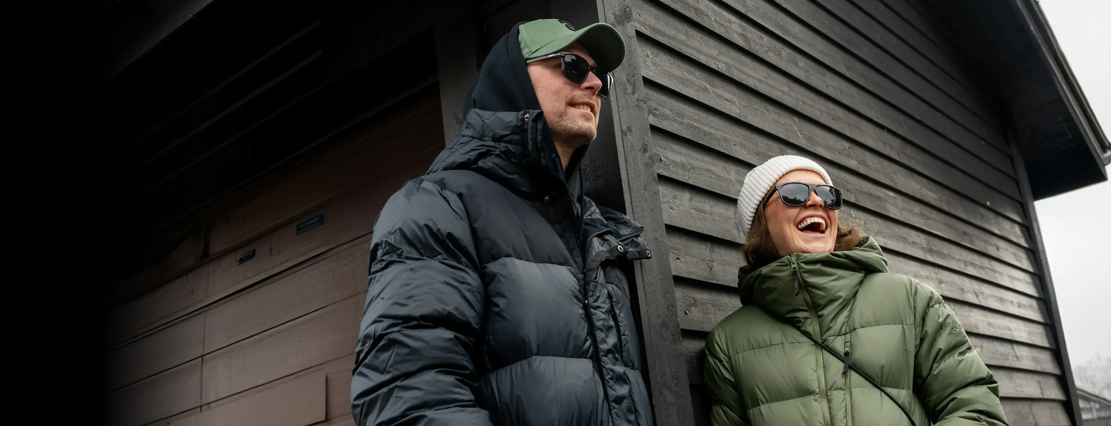 Two people in winter jackets and sunglasses smile and laugh as they stand against a dark wooden building.