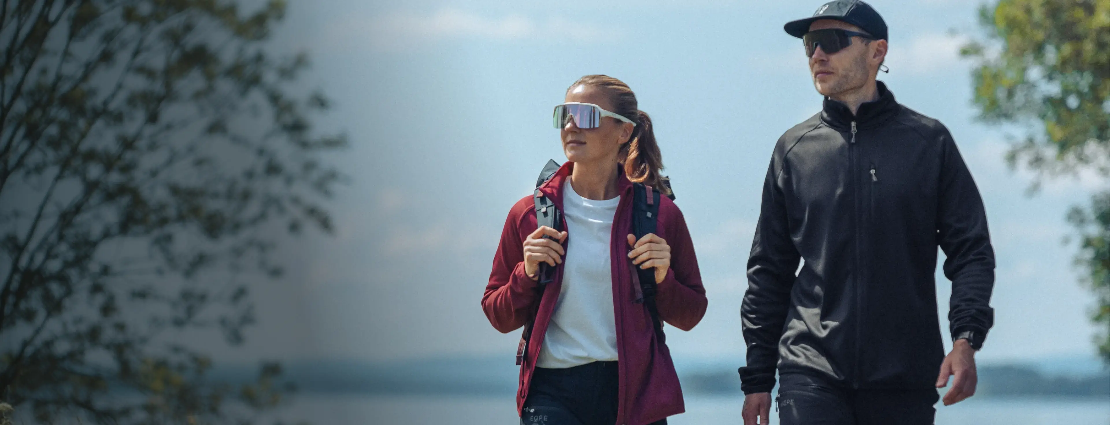 Two people in sunglasses and outdoor gear walk by a lake, surrounded by trees, on a sunny day.