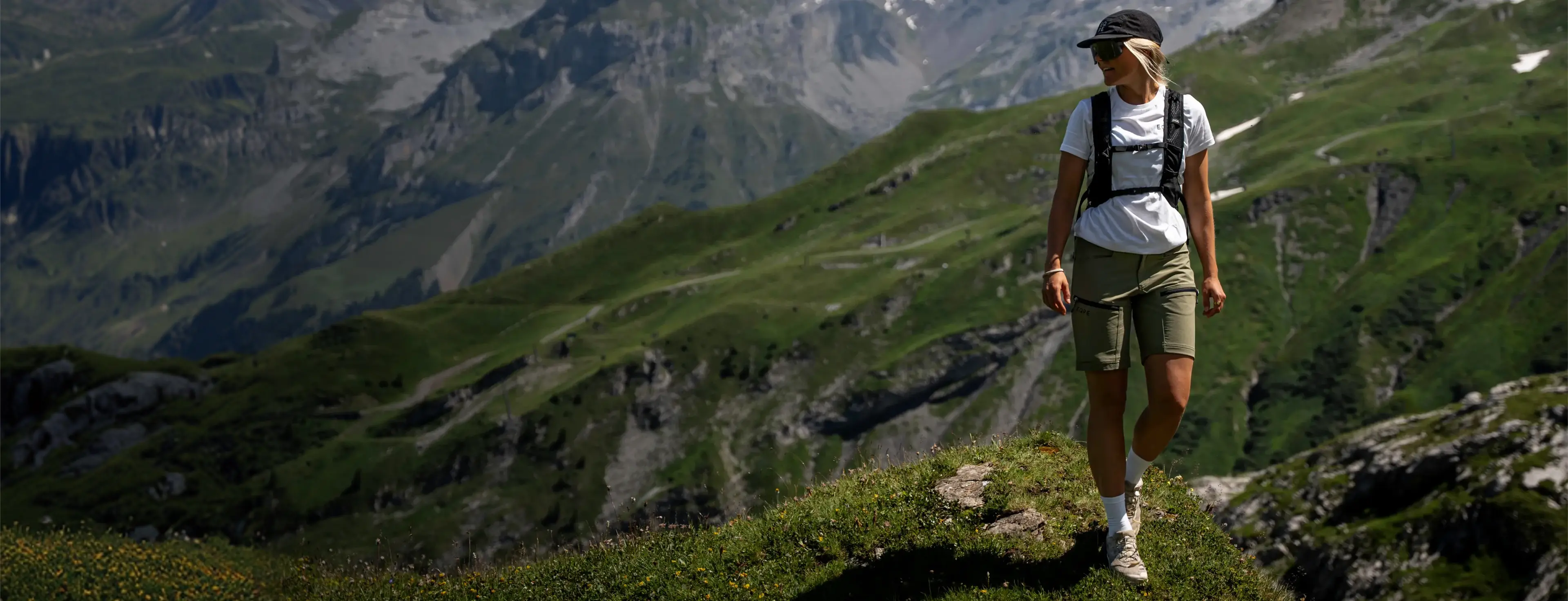 A person hikes along a grassy mountain ridge wearing a cap and backpack, with rocky peaks and lush valleys in the background.