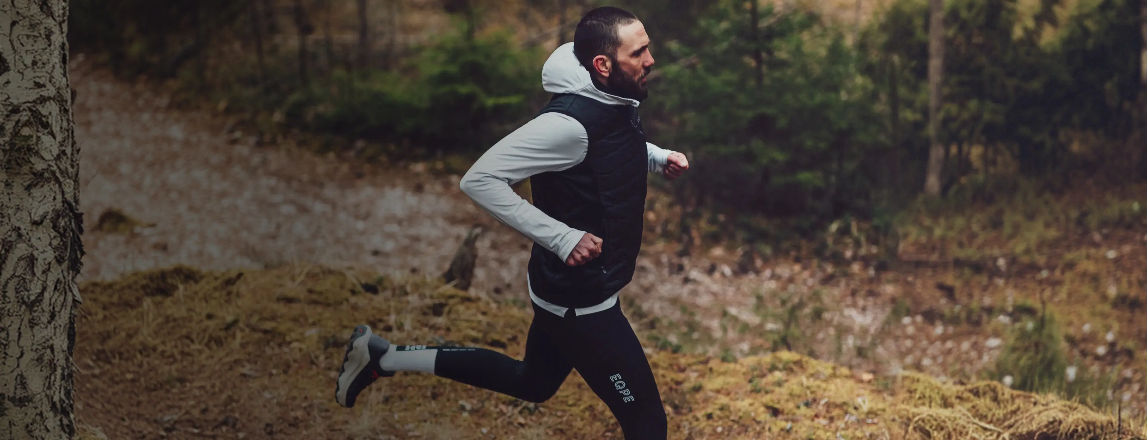 Man running through a forest path, wearing a black vest, white hoodie, and black leggings, surrounded by trees and fallen leaves.