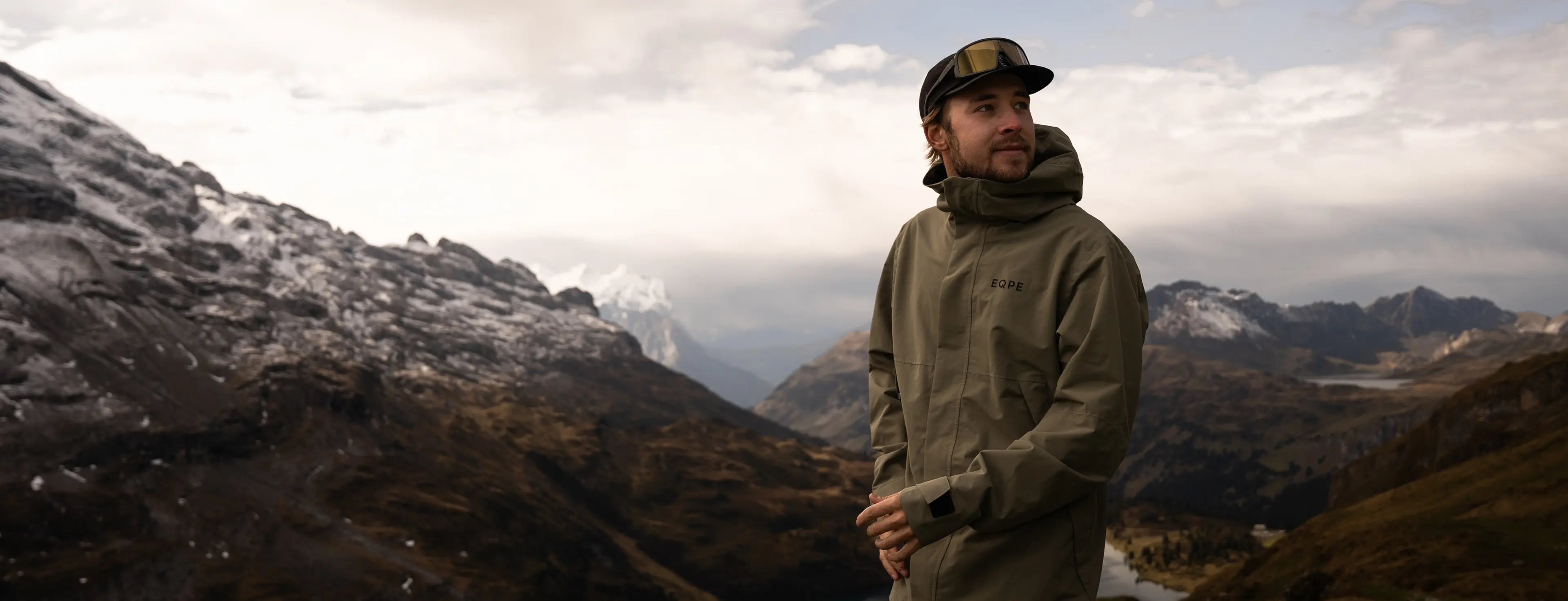 A person in a green jacket and cap stands in a mountainous landscape, with snow-capped peaks and a cloudy sky in the background.