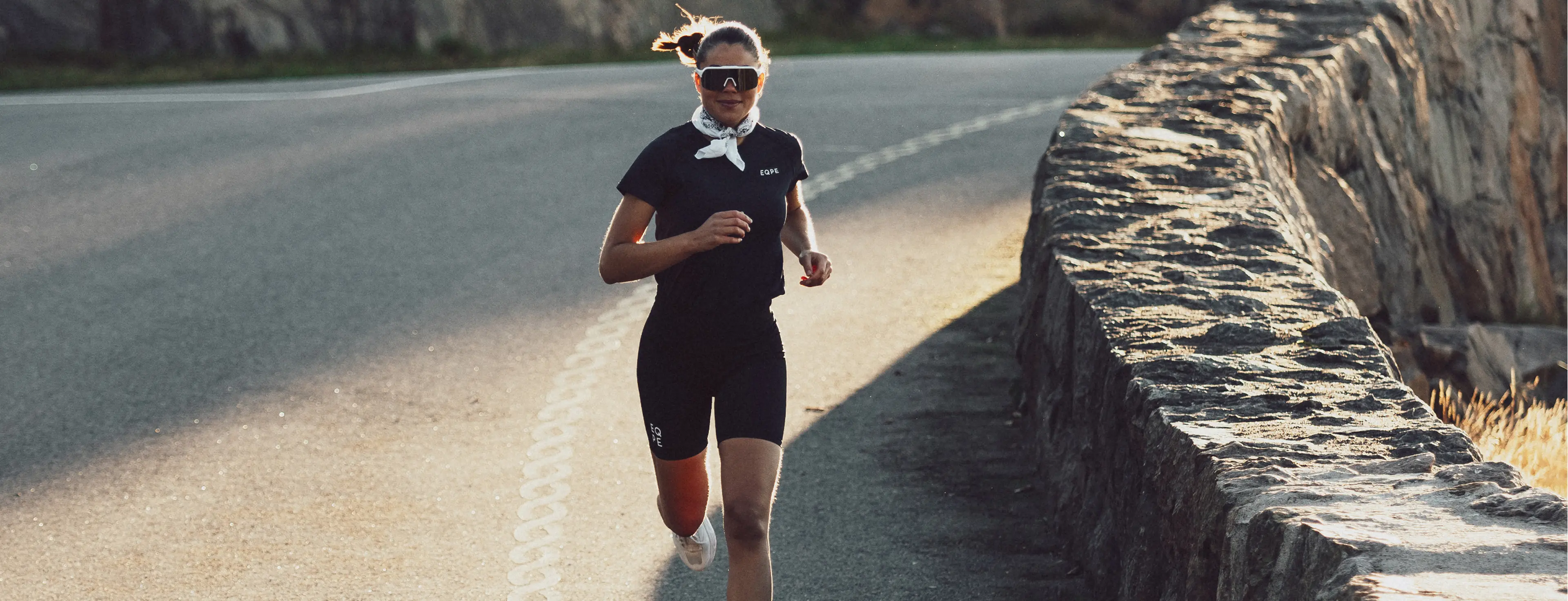 Woman running on a scenic road, wearing sunglasses, a white neckband, and a black outfit with "EQPE." Stone wall and sunlight in the background.