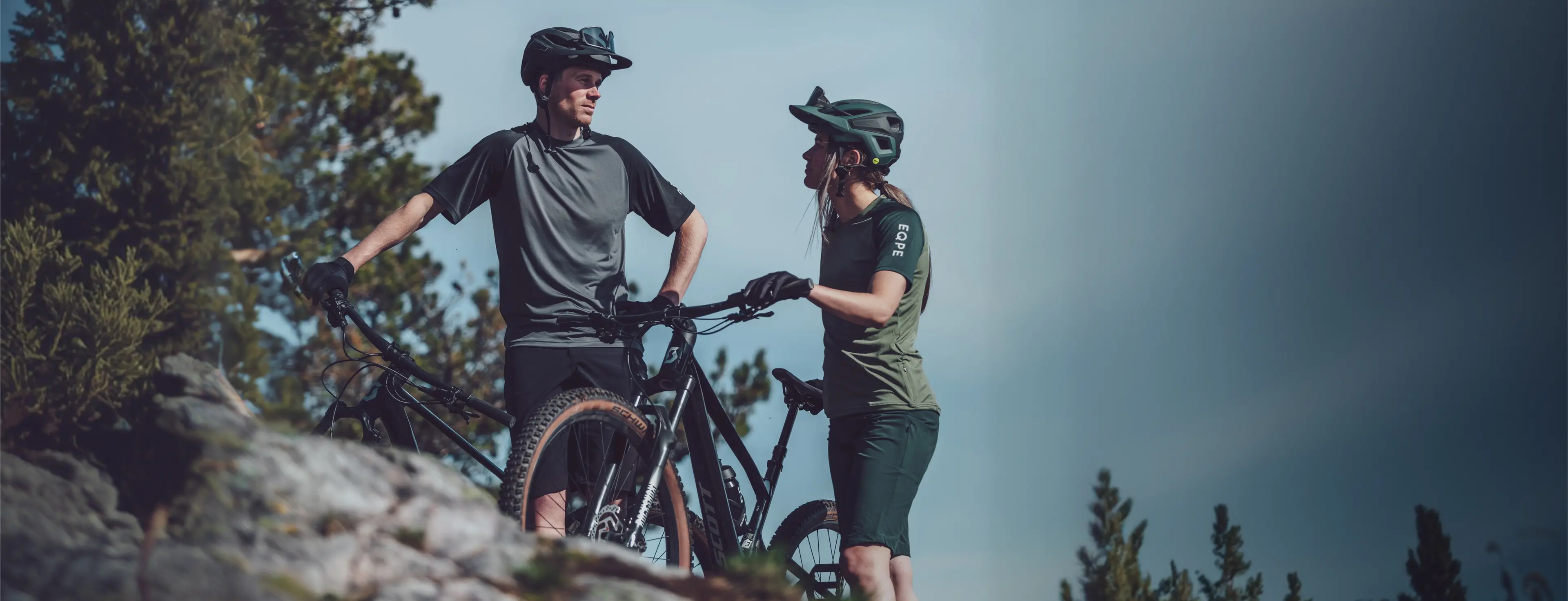 Two cyclists wearing helmets and gear, standing with their mountain bikes on a rocky trail, surrounded by trees and clear blue sky.