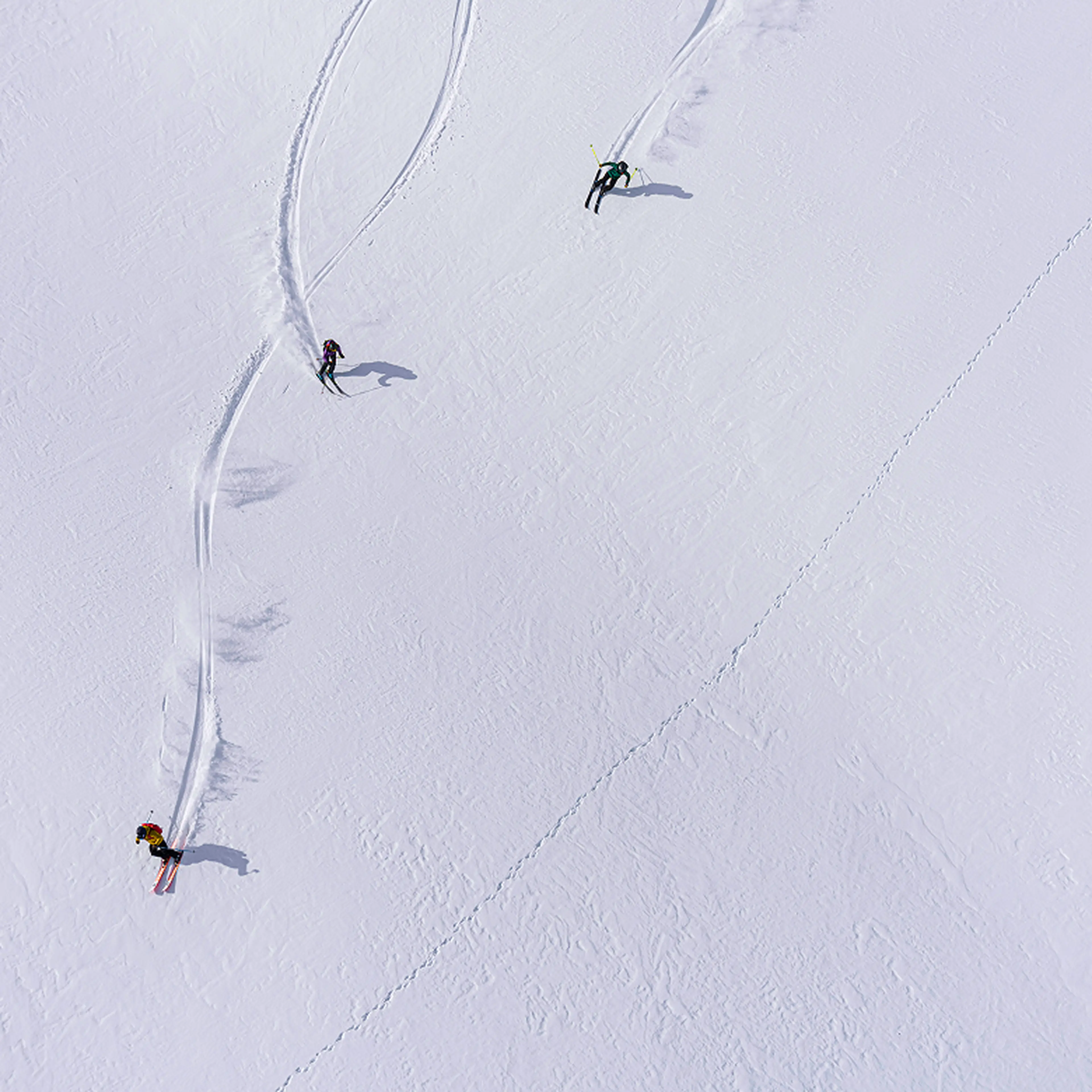 Three skiers leaving trails on a vast, untouched snowy slope, captured from above.