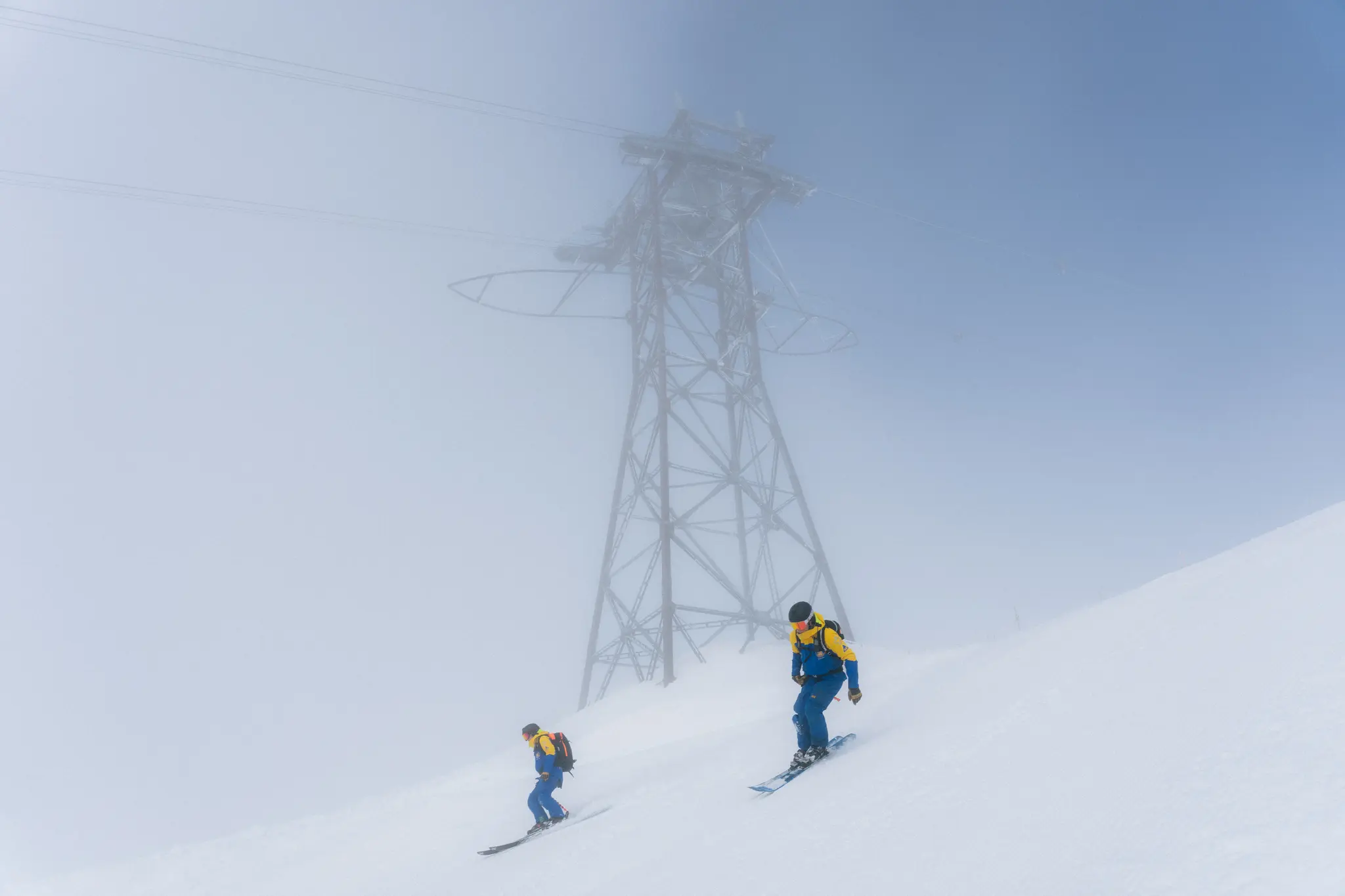 Two skiers in blue and yellow gear descend a snowy slope near a foggy metal tower on a misty day.