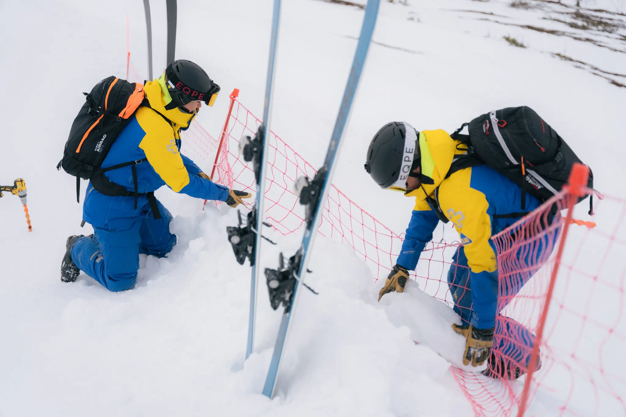 Two people in blue and yellow outfits work on a snowy slope, adjusting a red safety net.