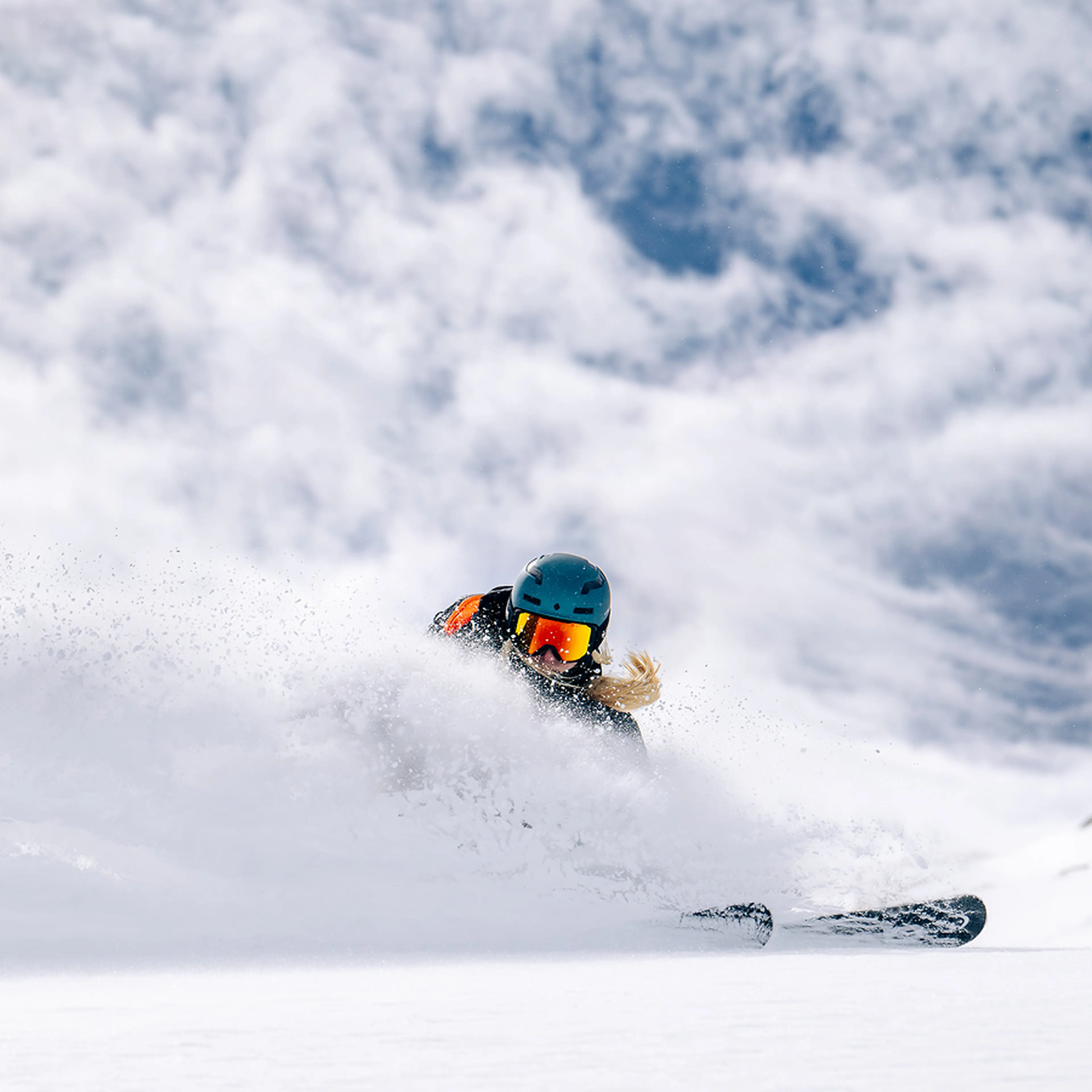 A skier in a helmet and goggles carves through fresh powder on a snowy mountain under a cloudy sky.