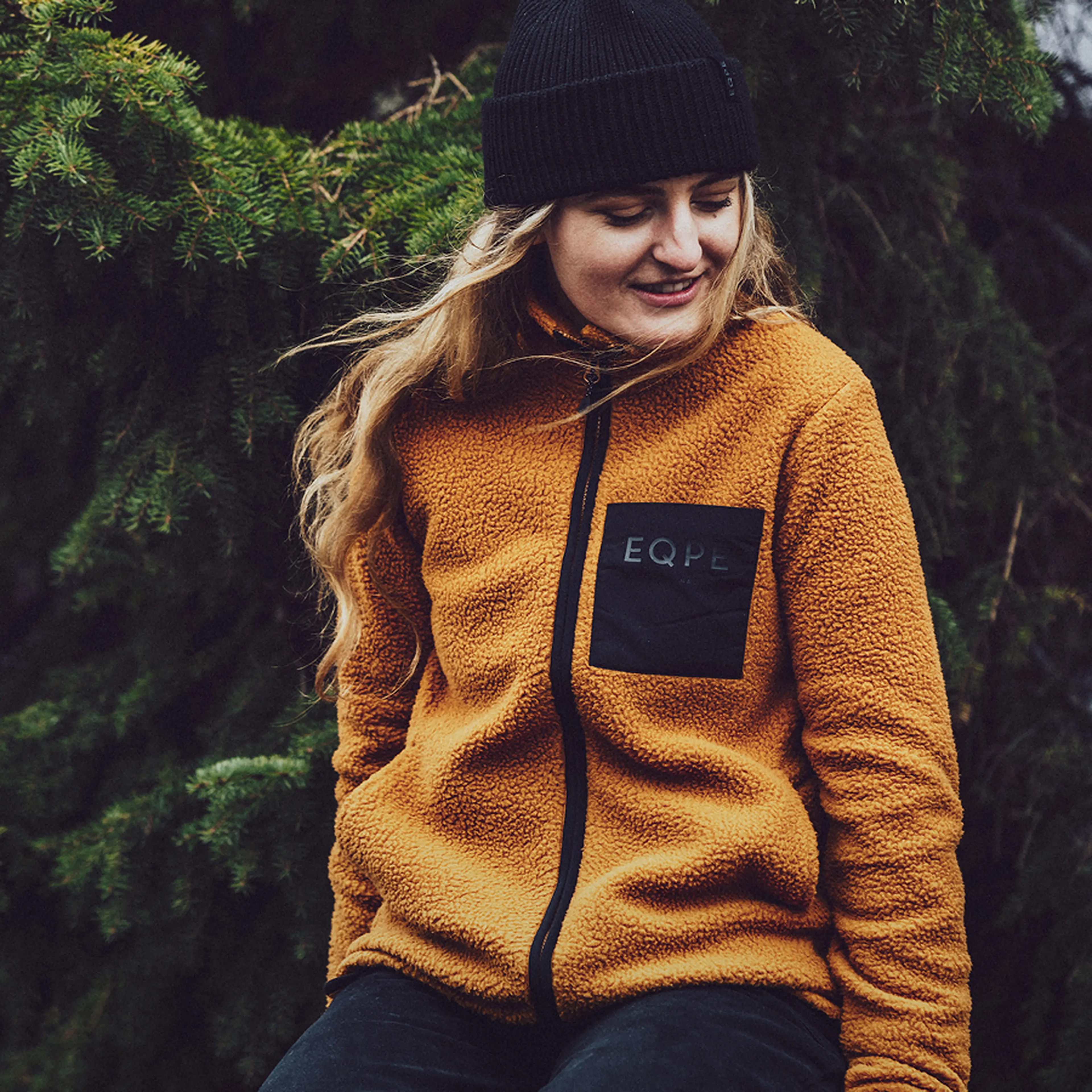 A person in a mustard-colored fleece jacket and black beanie sits in front of evergreen trees, smiling slightly and looking to the side.