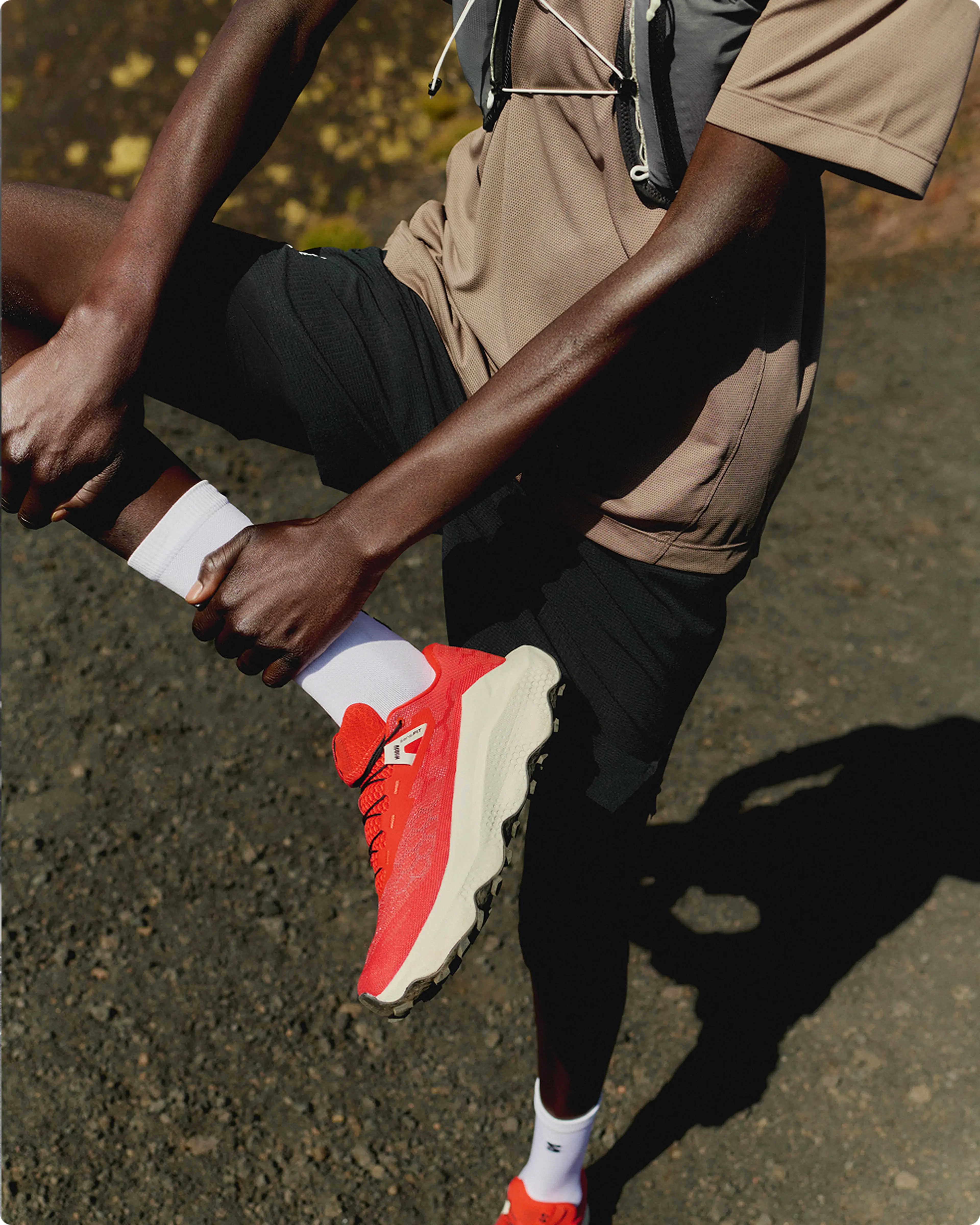 Person outdoors, wearing a brown shirt and black shorts, tying red sports shoes with white soles, and wearing a hydration vest.