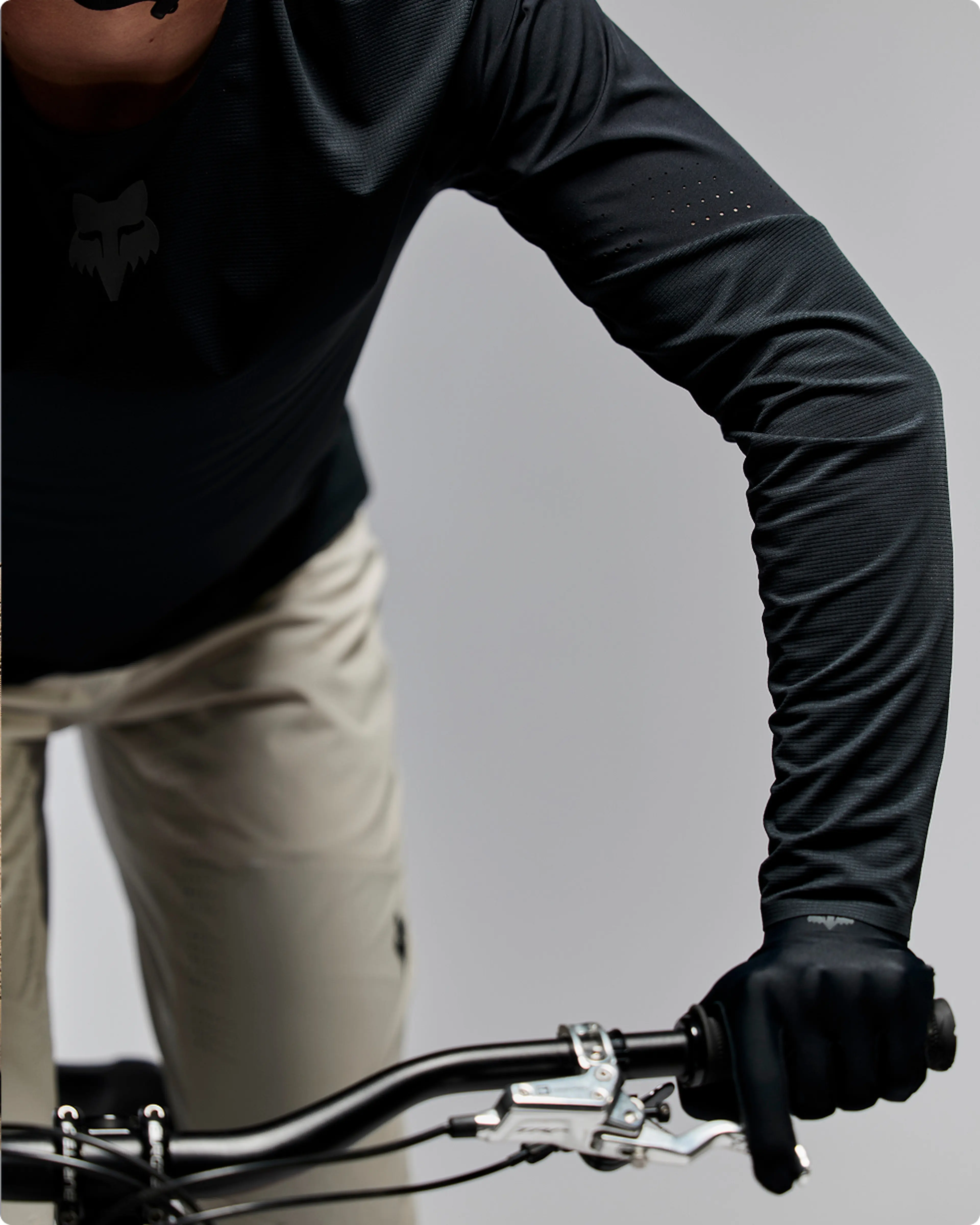 Close-up of a person in dark cycling gear and gloves holding mountain bike handlebars, wearing beige pants.