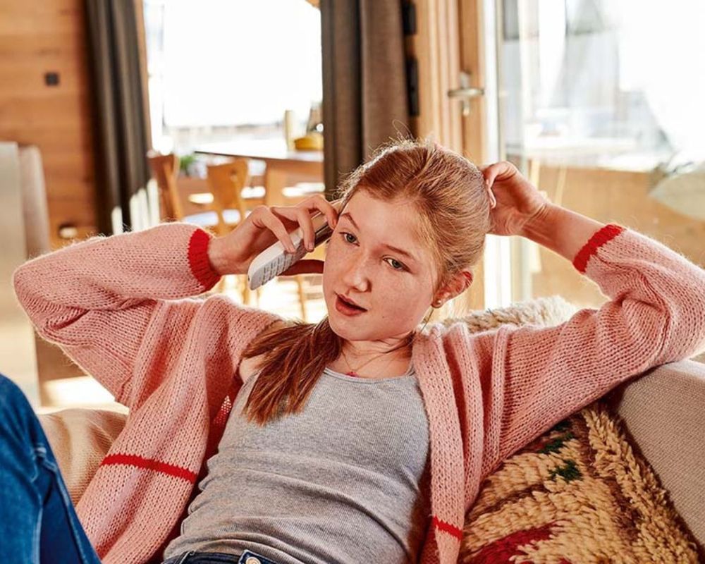 Dospievajúce dievča v ružovom svetri sa oddychuje na gauči a rozpráva sa cez bezdrôtový telefón, v pozadí je útulný domáci interiér.