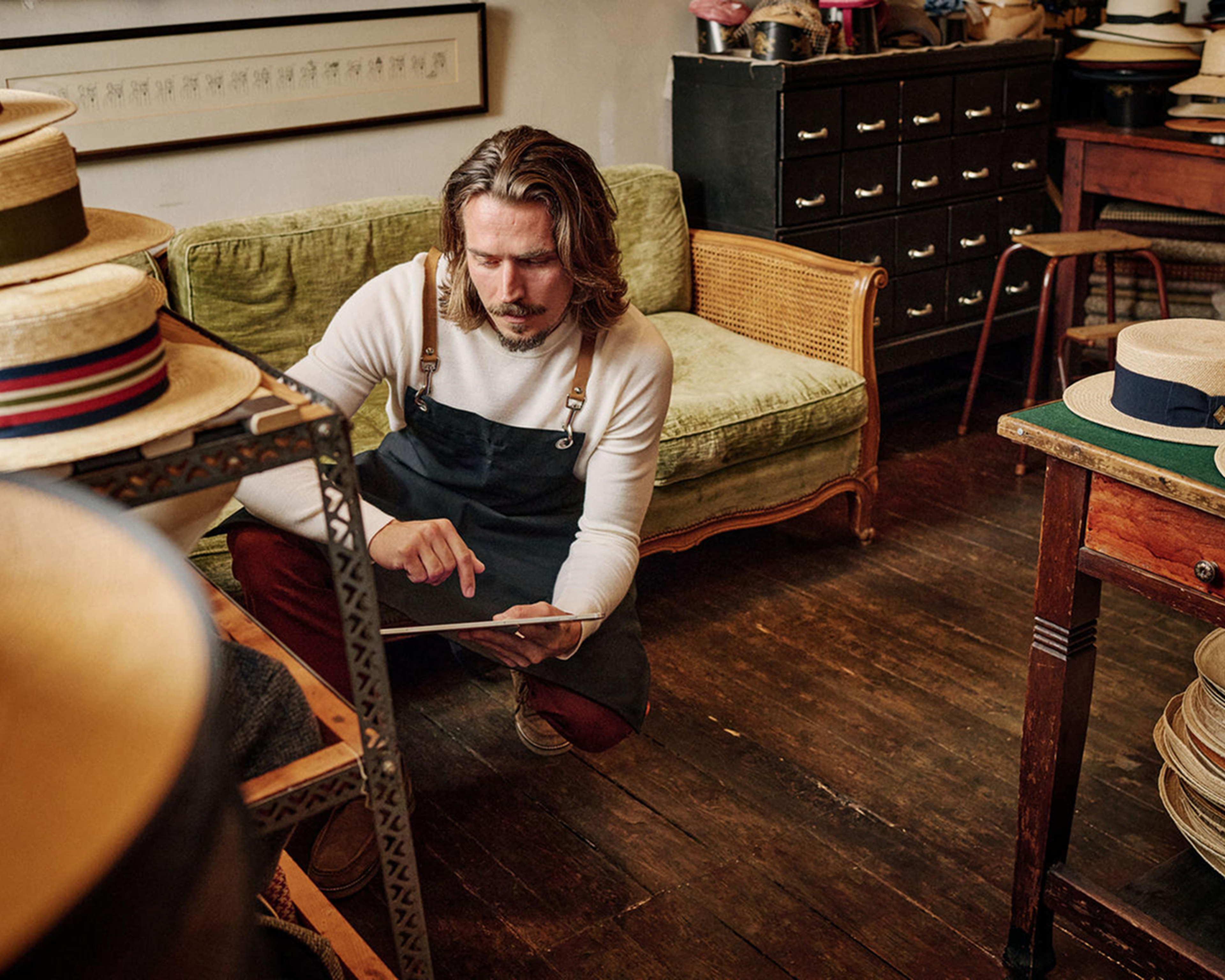 Muž v zástere kľačiaci na drevenej podlahe v miestnosti s klobúkmi. V ruke drží tablet.