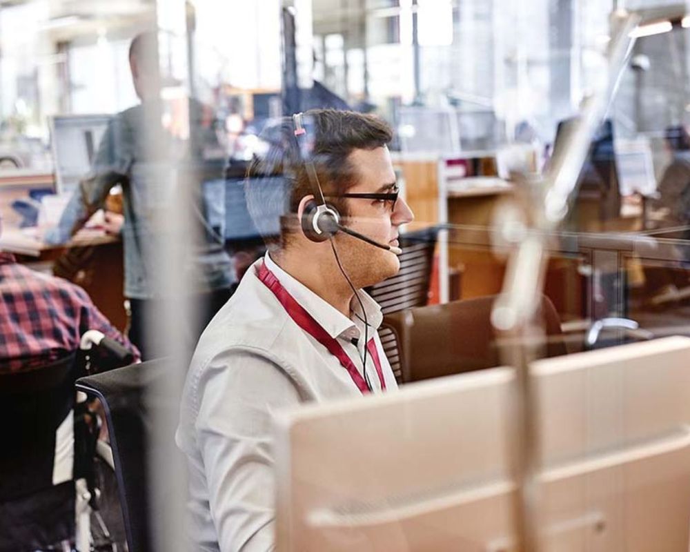 Muž s náhlavnou súpravou pracuje za počítačom v rušnom kancelárskom prostredí, videný cez sklenenú priečku.