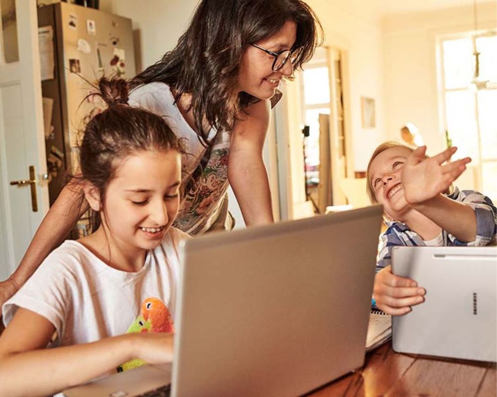 Žena sa usmieva na dve deti sediace pri stole s notebookmi, cez ktoré cez okno prúdi slnečné svetlo.