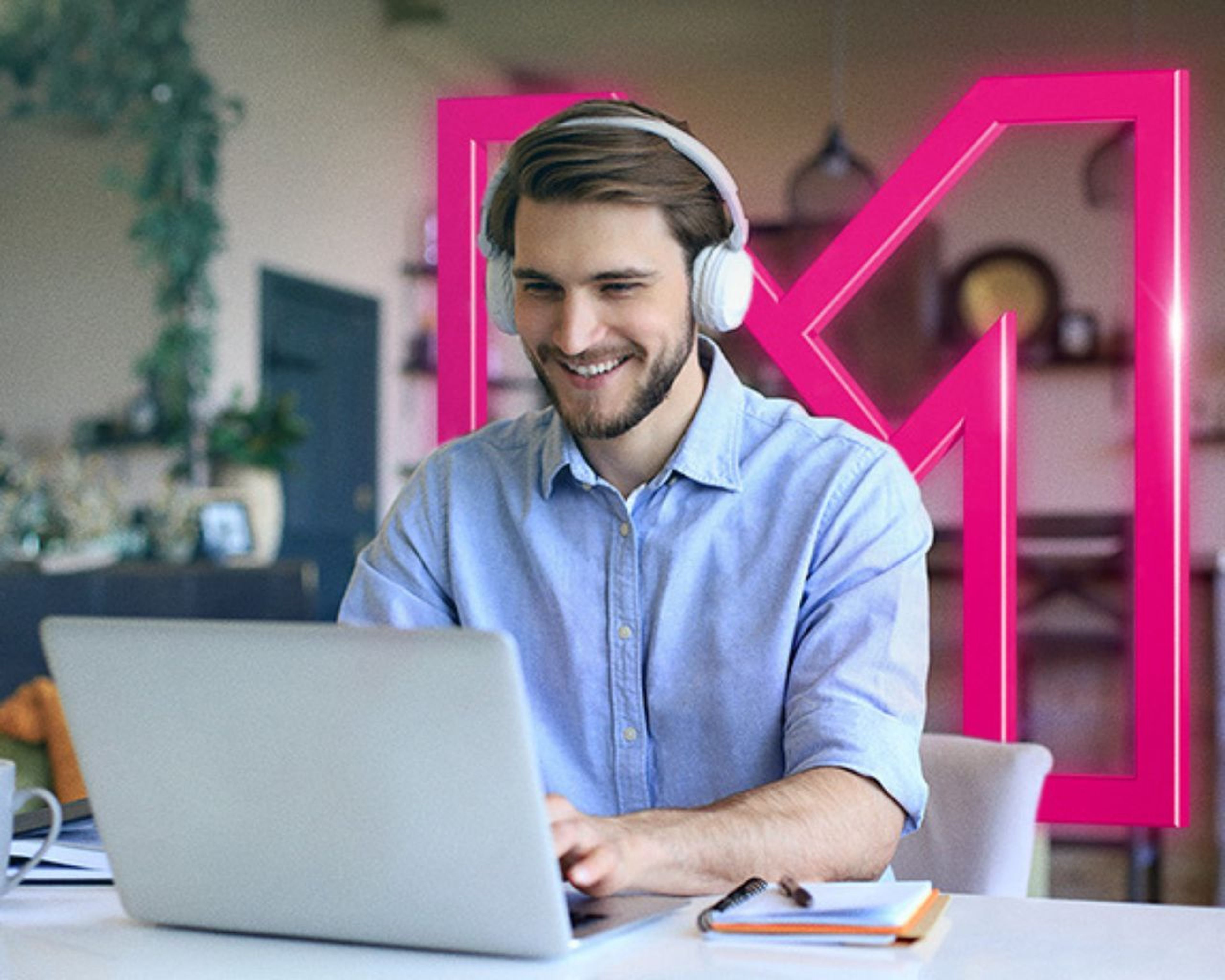 Muž v modrej košeli so slúchadlami na ušiach sa usmieva a používa notebook za stolom. V pozadí je veľké ružové „M“.