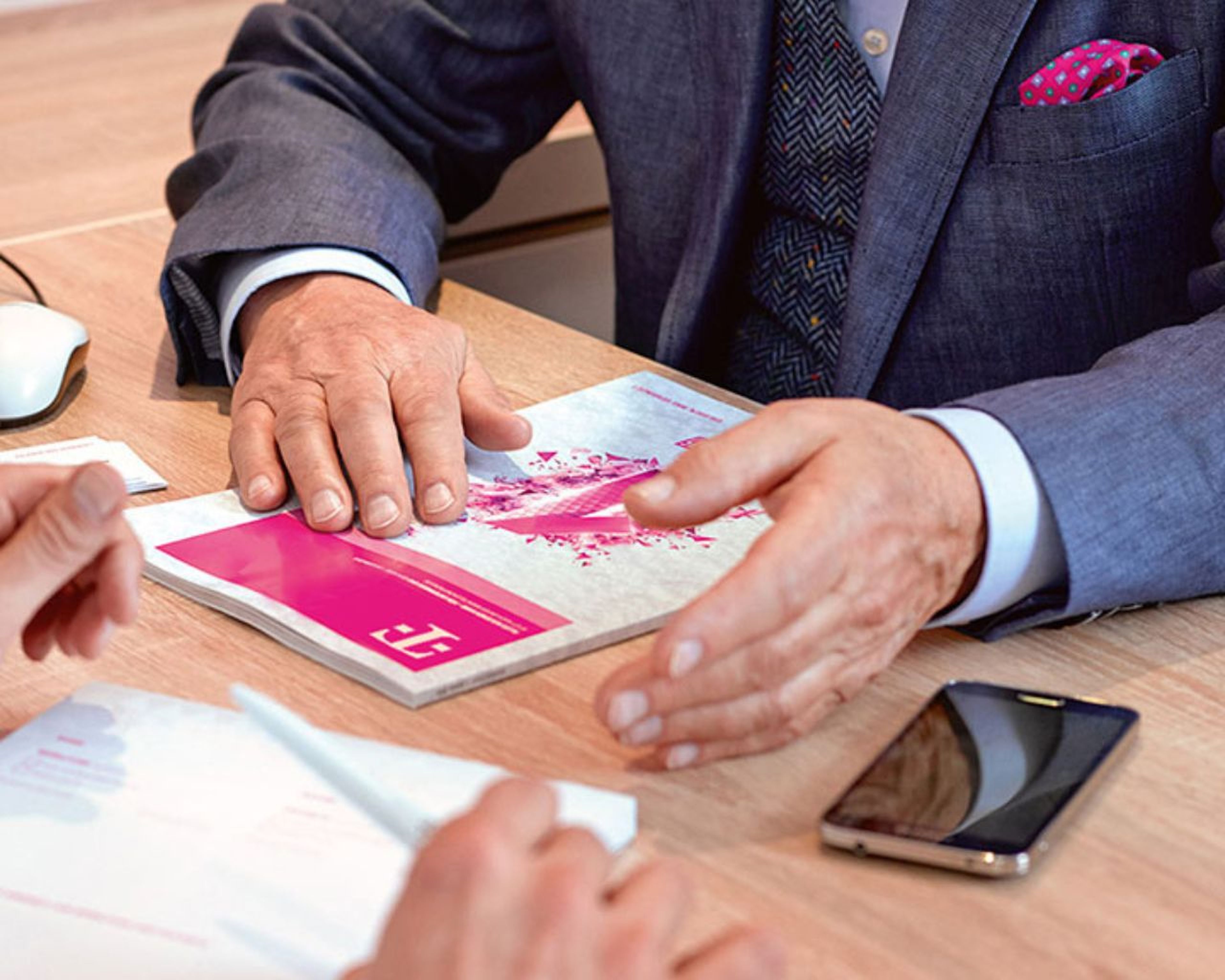 Naša silná podpora Dvaja ľudia na obchodnom stretnutí, jeden drží pero, druhý má na stole brožúru, vedľa seba smartfón a počítačovú myš.