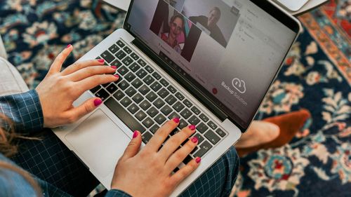 Osoba píše na notebooku počas videohovoru, na obrazovke sú viditeľní traja účastníci. Osoba má ružový lak na nechty.