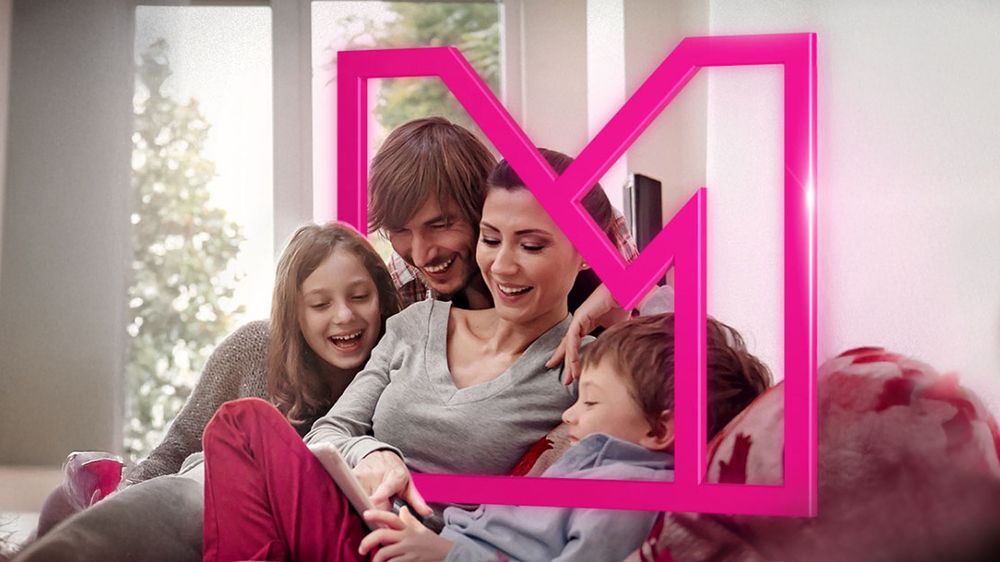 Usmievavá štvorčlenná rodina vrátane dvoch detí sedí spolu na gauči s veľkým ružovým logom „M“ na obrázku.