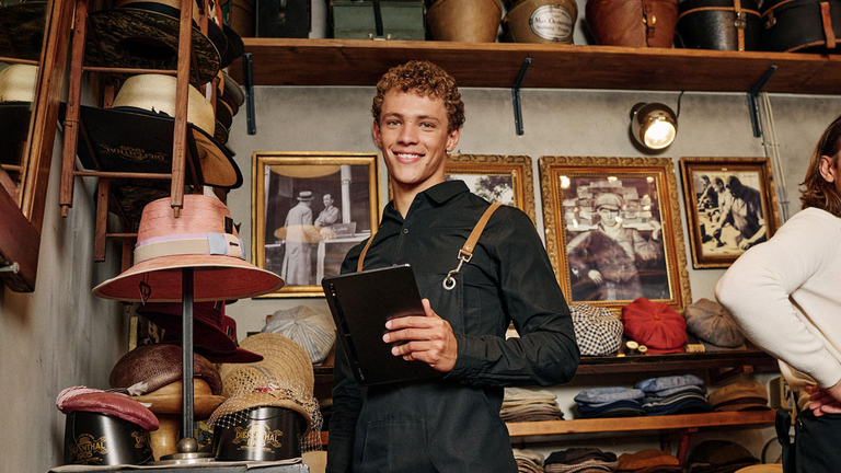 Mladý muž v obchode s klobúkmi držiaci tablet. Má oblečenú čiernu zásteru, obklopujú ho rôzne klobúky a zarámované fotografie na stene.
