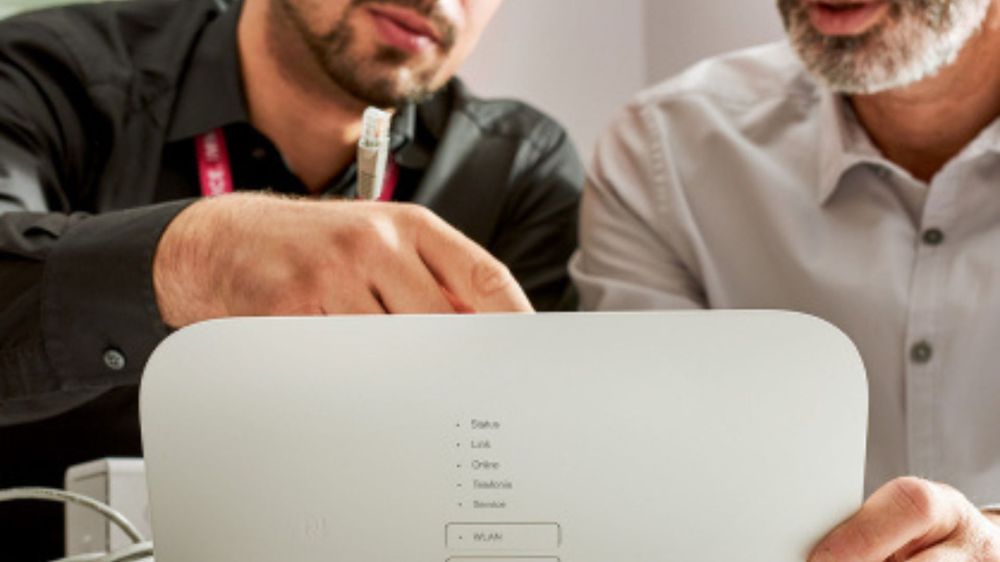 Dvaja muži diskutujú a ukazujú na biely Wi-Fi router na stole, neďaleko je notebook a káble.