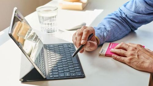 Osoba používajúca stylus na tablete s klávesnicou a držiaca knihu. Na stole je pohár vody a myš.
