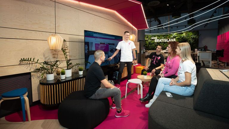 Skupina piatich ľudí v ležérnom oblečení sediacich a stojacich v modernej kancelárskej spoločenskej miestnosti s rastlinami, veľkou obrazovkou a nápisom „Ahoj Bratislava“.