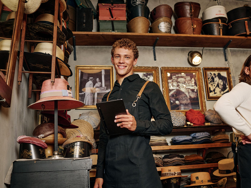 Mladý muž v obchode s klobúkmi držiaci tablet. Má oblečenú čiernu zásteru, obklopujú ho rôzne klobúky a zarámované fotografie na stene.