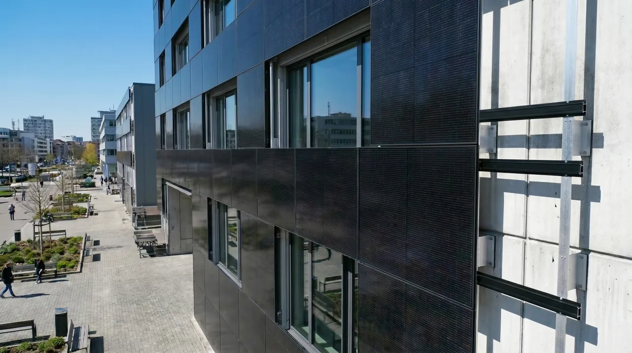 Modern building facade with dark solar panels, city street view with trees, benches, and pedestrians under a clear blue sky.