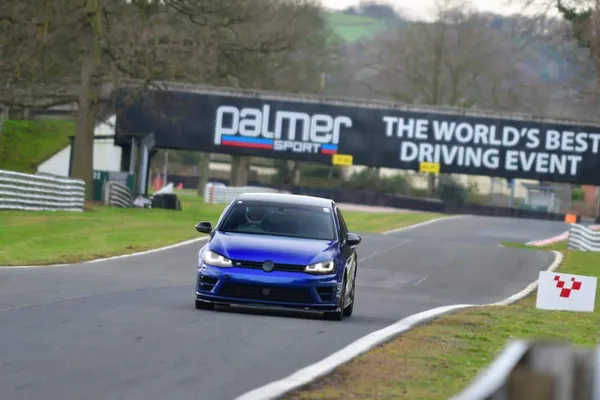 A blue car drives on a racetrack under a banner reading "Palmer Sport: The World's Best Driving Event," surrounded by trees and greenery.