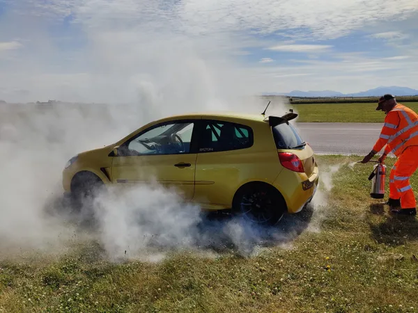 Yellow car with smoke around it on grass; a person in an orange outfit approaches with a fire extinguisher.