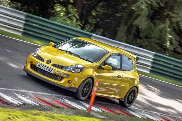 Yellow sports car taking a sharp turn on a racetrack, surrounded by green and white barriers, with trees in the background.
