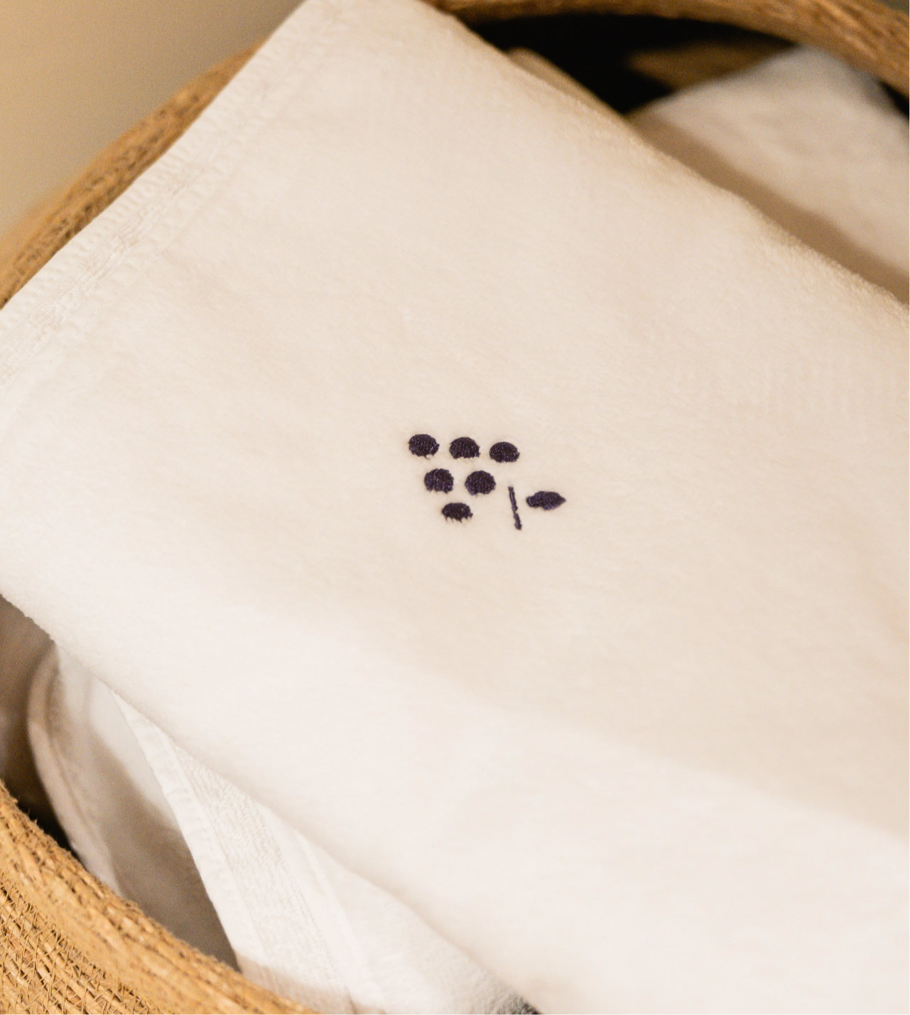 0074_20260121_MOREL__MG_4236_HD_Caudalie@2x White towel with a small blue embroidered grape design folded in a wicker basket.
