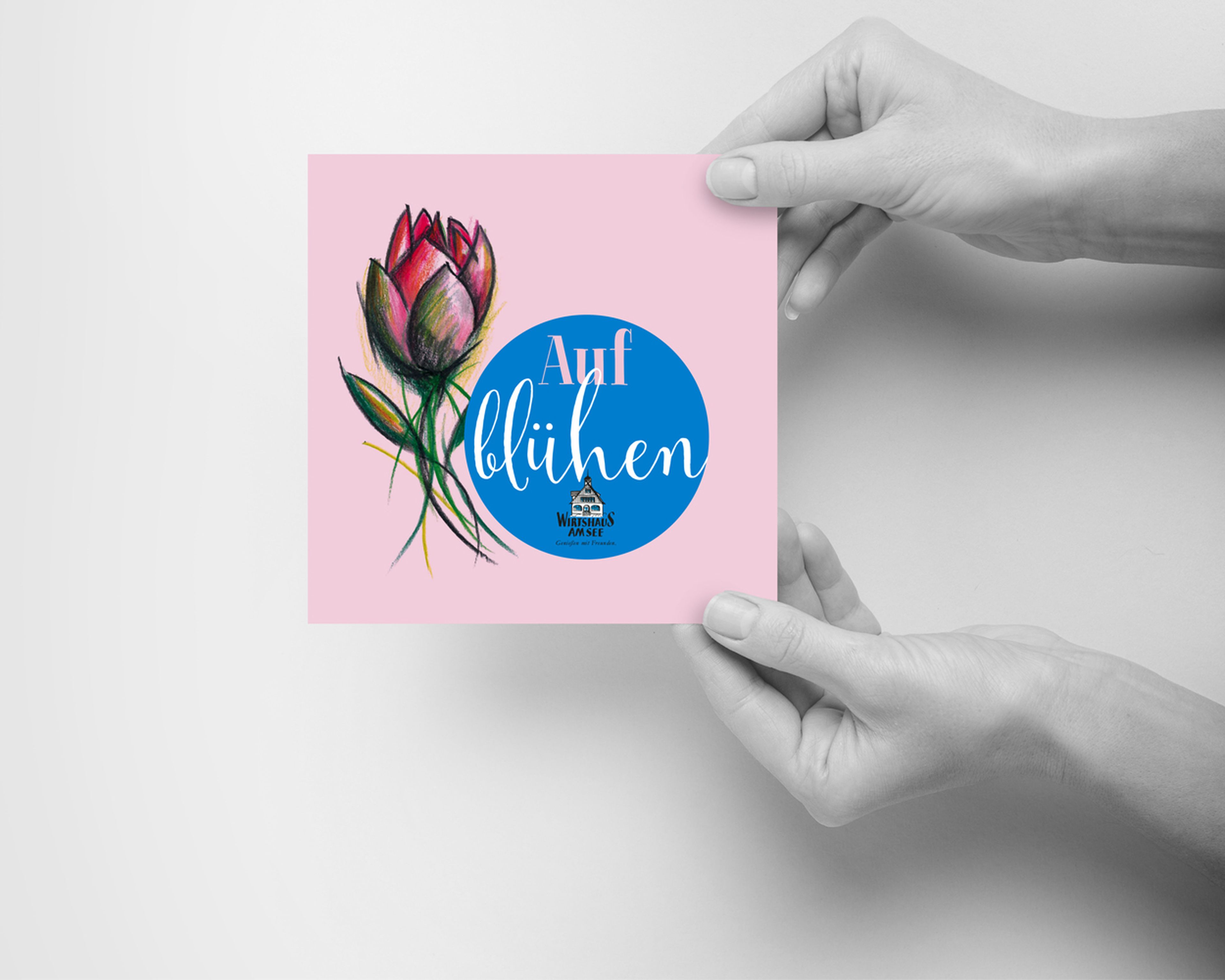 Hände halten eine rosa Karte mit einer roten Blumenillustration und dem deutschen Text „Auf blühen“ in einem blauen Kreis. Mailing Wirtshaus am See, Bregenz – Gestaltung: Ölz GrafikDesign