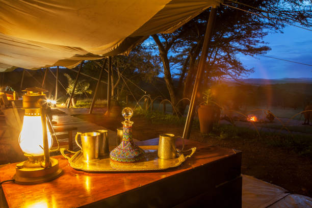 A cozy outdoor setting at dusk with a lantern-lit table, ornate decanter, and mugs. A campfire glows softly in the background.